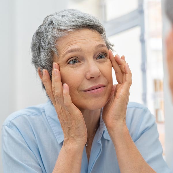 Older woman looking in the mirror examining her upper face area around her eyes and thinking about facial plastic surgery.