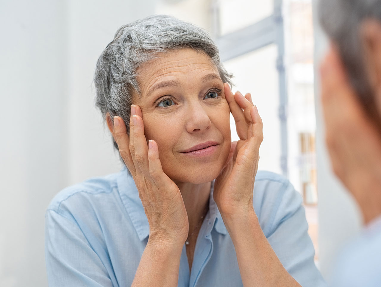 Older woman looking in the mirror examining her upper face area around her eyes and considering blepharoplasty.